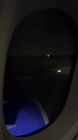 This video shows a view from an airplane window at night. Outside, the runway lights are visible, with a red light in the distance and several white lights closer to the plane. The ground appears to be dark and possibly grassy. A portion of the airplane's wing or engine, which is painted blue, is visible at the bottom of the frame. The sky is dark and appears to be overcast. The video captures the plane moving, as indicated by the slight blur of the lights and the changing perspective.