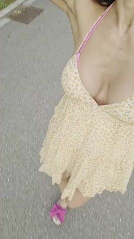 The video shows a close-up, high-angle shot of a woman walking outdoors. She is wearing a short, floral-patterned dress with pink straps and pink sandals. The camera is positioned above and in front of her, capturing her from the chest down. The background appears to be a paved surface, possibly a sidewalk or road. The woman's hair is dark and falls over her shoulders. She is looking down, and her right arm is bent at the elbow, with her hand near her head. The overall impression is that this is a candid shot, possibly taken by the woman herself or someone walking with her.