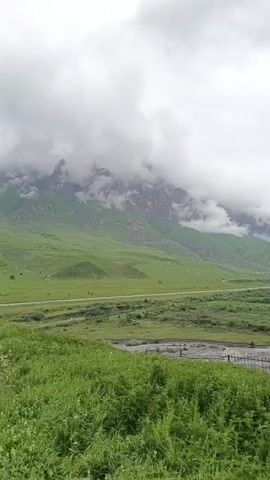 A panoramic view captures a verdant valley cradled between majestic, cloud-shrouded mountains. A winding river snakes through the valley floor, its banks lined with sparse vegetation. In the foreground, lush green grass grows densely, obscuring the lower portion of the frame. A road can be seen in the middle ground, curving along the valley. The sky is overcast with heavy clouds, lending a dramatic and moody atmosphere to the scene. The camera pans slowly from left to right, revealing the vastness of the landscape.