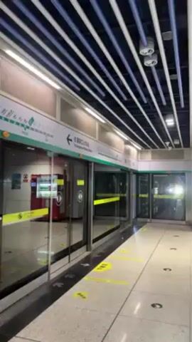 The video shows the interior of a subway station. The camera is positioned facing a set of sliding glass doors that lead to the train platform. The platform is visible through the doors, and a train can be seen approaching. The doors are flanked by signs indicating the name of the station in both Chinese and English. The floor is tiled and has yellow markings indicating where people should stand. The ceiling is decorated with parallel white and blue stripes. There is a vending machine visible behind one of the glass doors.
