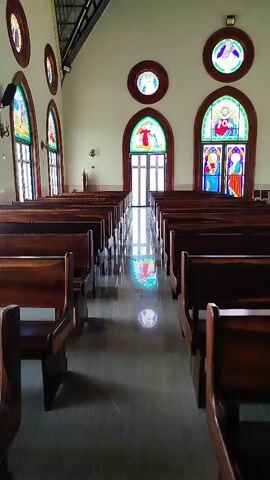 The video shows the interior of a church. Rows of wooden pews face a central aisle that leads to a set of double doors at the far end. Stained-glass windows with religious imagery are visible on the walls, and their reflections can be seen on the polished floor. The ceiling is high and appears to be made of metal. There are no people or animals visible in the video, and no actions or interactions are depicted. The environment is calm and still.