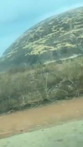 The video shows a view from inside a moving vehicle, looking out towards a rocky hillside. The hillside is covered in sparse vegetation, mainly grasses and small shrubs. The sky is overcast and a light bluish-green color, possibly due to the window tint. The road is visible in the lower right corner of the frame, with a brownish-red surface. The camera appears to be shaky, likely due to the movement of the vehicle.