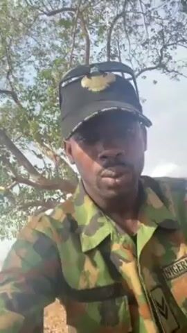 A man in a Nigerian Army uniform and a black baseball cap is recording a video of himself. He appears to be in a rural or semi-rural environment, as he is seen against backdrops including trees, dirt roads, and a glimpse of a paved road with buildings in the distance. The sky is overcast, and the light is diffuse. The man is looking directly at the camera, and his expression is serious. The video appears to be taken from a relatively close range, focusing primarily on the man's face and upper body.