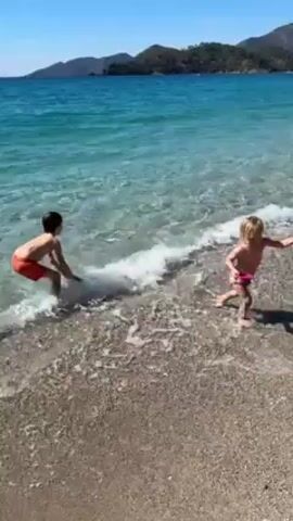 The video shows two young children playing in the surf on a beach. The water is clear and turquoise, and the sand is a mix of tan and grey. The beach slopes up toward a hillside covered in green trees.  The sky is blue and cloudless.  One child, a boy in red swim trunks, is walking deeper into the water while holding a small white bucket.  A second child, a girl in a pink swimsuit, is walking along the shoreline, splashing in the shallow waves.  The camera pans slightly to follow the children's movement.  The overall impression is that it is a bright, sunny day at a scenic beach.