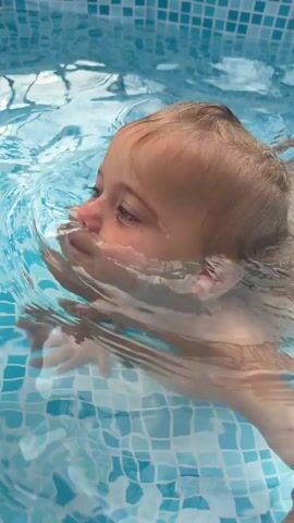 A child with blonde hair is partially submerged in a tiled swimming pool. The child's face is visible, and their head is tilted to the side. A person's arm is supporting the child. In the background, another person with dark hair and a beard is visible, smiling and looking at the child. The environment appears to be outdoors, with a fence and trees in the distance. The water is blue and clear, with ripples and reflections on the surface.