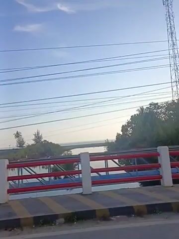 The video shows a view from a moving vehicle, likely a car, looking out over a body of water. The foreground features a bridge railing with white posts and red and blue horizontal bars. The road surface in front of the railing has yellow and black diagonal stripes. In the background, there is a calm body of water, possibly a river or a bay, with trees and vegetation along the shoreline. The sky is blue with some wispy clouds, and there are several overhead wires visible. The overall impression is of a peaceful daytime scene from a transit route.