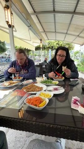A man and a woman are seen eating a meal at an outdoor restaurant in Finike, Turkey. They are seated at a dark-colored table with a glass top, under a white canopy with overhead lamps.

The man, on the left, wears a blue hoodie with "SUPER WET" printed on it and a multi-colored scarf. He is eating from a plate containing what appears to be eggs or cheese with a side salad. The woman, on the right, wears glasses and a dark jacket, and is actively eating from her plate, which holds a salad and other ingredients, using a fork.

On the table in front of them are several dishes: a large, flat dish with a brownish, crumbly food (possibly a ground meat dish or kebab), a bowl of shredded carrots with a lemon wedge, a bowl of small yellow items (likely pickled vegetables or fruit), and a small plate with a purple-colored dish (possibly a beetroot salad or dip). A mobile phone and tissue packets are also visible on the table.

In the background, other diners are visible at another table, along with lush greenery and distant buildings, creating a pleasant outdoor dining atmosphere. Both individuals appear relaxed and focused on their meal. The audio indicates they are speaking in Russian.