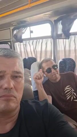 The video shows a selfie taken from inside a bus. In the foreground, a man with graying hair and a black t-shirt looks directly at the camera. Behind him, to the right, another man wearing sunglasses and a brown t-shirt with a white design on it is visible. He is looking to his left and appears to be speaking or gesturing. Further back in the bus, several other passengers are seated. The environment is typical of a bus interior, with windows covered by curtains and overhead luggage compartments. The lighting suggests it is daytime.