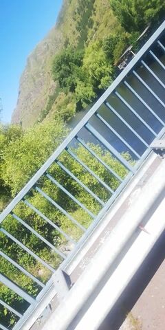 The video shows a view from a bridge over a river. Green trees and vegetation are visible on the banks of the river and on the hillside. A mountain is visible in the distance. The sky is blue and clear. The bridge has a metal railing. The video is shaky, suggesting it was filmed while walking or moving.