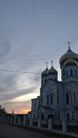 The video shows a large, ornate Orthodox church with multiple golden domes and crosses on top. The church is white and blue, with gold accents around the windows and architectural details. A black metal fence surrounds the church grounds. Power lines are visible against the sky. The sky is a mix of blue and gray clouds, with a hint of orange sunset visible in the distance. The camera pans slightly upward and across the church's facade, highlighting its architectural features. The video appears to have been taken at dusk or dawn.