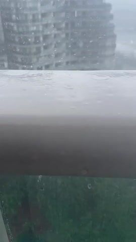 Raindrops are hitting a railing. In the background, there is a blurry building and trees. The sky is overcast.