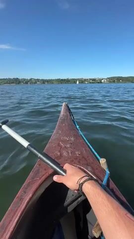 The video shows a first-person view from a canoe on a body of water on a sunny day. The camera is positioned at the front of the canoe, focusing on the bow and the water ahead. A hand holding a paddle rests on the side of the canoe. The canoe is reddish-brown and has some writing on the side. The paddle has a silver shaft with a black handle. The water is dark and has small waves. In the distance, there is a shoreline with trees and some buildings. Two sailboats are visible on the water. The sky is blue with some scattered clouds.