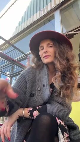 A woman with long, wavy brown hair and a red hat is sitting outside in front of a building. She is wearing a grey knitted cardigan over a black floral dress and black tights. She is holding a light green mug with a tan rim and drinking from it. The background shows the side of a building with a corrugated metal roof and a window. There are metal shelves and some plants visible.