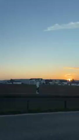 The video shows a moving view from inside a car, likely the passenger seat, looking out toward the right. The scene depicts a rural landscape at sunrise, with houses and trees visible in the distance under a blue and orange sky. A highway barrier runs along the bottom of the frame. A car side mirror is visible in the lower right corner. The car appears to be moving at a moderate speed.