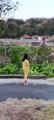 A young person in a yellow tracksuit stands facing away from the camera, looking at a village nestled in a hillside. The village features houses with red tile roofs. The young person then begins to walk away down a paved road that curves to the right. The camera follows, panning slightly to the right and upward to show more of the village and the surrounding hills. The sky is cloudy.