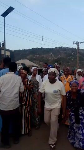 The video shows a group of people, mostly women, walking together outdoors. Many of the women are wearing colorful, patterned dresses and headscarves. One woman is wearing a white t-shirt and khaki pants.  The group is walking on a paved road or street. In the background, there is a hill or mountain, buildings, and utility poles with wires.  The sky is overcast. The camera appears to be hand-held and moves along with the group.  Some of the people are talking and smiling.