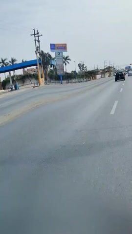 The video shows a daytime scene from a moving vehicle on a paved road. The road has at least two lanes in the direction of travel, marked by a dashed white line. Other vehicles, including a black SUV/hatchback, are seen ahead. To the left, there's a side road or turning lane, with a large blue and white sign prominent in the foreground. This sign reads "CASAS EN MOCHE" (Houses in Moche), followed by a phone number "994334640" and additional text like "LOTES AL CLUB" and partially visible words "LUGARES, MARE...". Beyond the sign, there are power poles with electrical lines, sparse greenery, and several palm trees, indicating a warm climate. The sky is clear and bright. The scene suggests an urban or semi-urban environment in Moche, Peru.