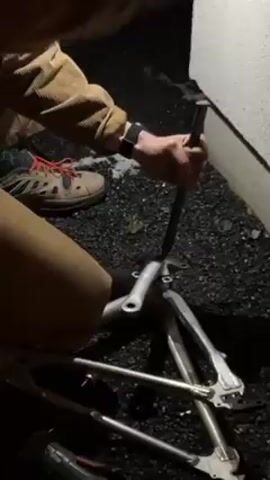 A person is kneeling outdoors, next to a concrete wall, working on a silver bicycle frame that is lying on dark gravel. The person is wearing a brown jacket, tan pants, a watch, and Salomon brand shoes. They are using a long black metal rod to hammer on a component of the bike frame. After hammering, the person moves the frame. The lighting is dark, making it hard to see details.