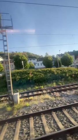 The video shows a view from a moving train. Train tracks are visible in the foreground and middle ground. To the right of the tracks is a tall signal post with lights. In the background, there are trees, bushes, and some buildings on a hillside. The sky is blue with some white streaks of light. The lighting suggests it is daytime, possibly morning or late afternoon.