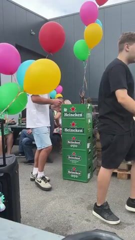 The video opens with a man in a white t-shirt and blue shorts standing in front of a stack of green Heineken beer cases. He is holding a bunch of colorful balloons, including pink, yellow, green, and blue ones. In the background, there are people sitting at tables, and another man is seen interacting with the first man. The setting appears to be an outdoor gathering, possibly a party or event, with a modern building in the background. The ground is paved, and there are some wooden pallets with items on them. As the video progresses, another person in a black t-shirt and shorts approaches, and the two men appear to be moving the beer cases. More balloons are visible in the background, adding to the festive atmosphere. The focus remains on the interactions between the people and the presence of the beer and balloons.