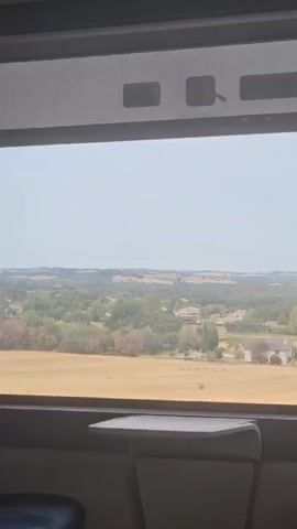 This is a first-person video taken from inside a train, looking out the window at a rural landscape. The train appears to be moving at a moderate speed, as the scenery outside is blurred and shifting. The window is large, with a tray table attached to the seat in front. The landscape consists of rolling hills covered in fields of dry, golden grass and scattered trees. In the distance, there are small clusters of houses and buildings, suggesting a rural village or scattered settlements. The sky is overcast with a pale blue hue. The video is brief, lasting only a few seconds, and offers a glimpse of a peaceful, bucolic scene experienced during train travel.