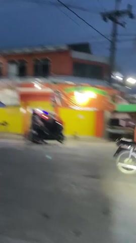 The video begins with a view of a street at dusk or dawn. The sky is a deep blue, and streetlights illuminate the road. On the left side of the street, there are buildings with signs, one of which appears to be an advertisement with orange and yellow colors. The road is paved and appears to be mostly empty, with a few vehicles visible in the distance.

As the video progresses, the perspective shifts to show a person riding a motorcycle. The rider is wearing a helmet and is on a scooter-like motorcycle. In the foreground, the handlebars and part of the motorcycle's instrument panel are visible. The video captures the rider's point of view as they navigate the street, passing other motorcycles and buildings. Several other motorcycles are seen on the road, some with riders and headlights on. Trees and utility poles with wires line the street, contributing to the urban or semi-urban environment. The lighting suggests it is either early morning or late evening. The overall atmosphere is calm and ordinary, depicting a typical street scene with motorcycle traffic.