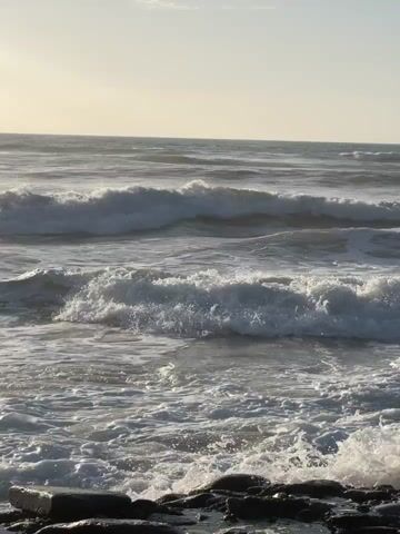 A video shows a beach with waves crashing against the shore under a bright sky. The ocean is vast, with several lines of waves rolling in towards the viewer. The waves nearest to the camera are white with foam as they break and recede. Further out, the waves are darker, showing the movement of the water. The sky is bright and hazy, indicating either early morning or late afternoon, with the sun casting a soft light over the scene. The horizon is visible as a distinct line between the ocean and the sky. The foreground includes some dark, wet rocks or debris at the very bottom of the frame, partially submerged in the water. The overall impression is one of natural beauty and movement.