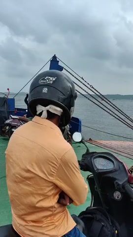 A man wearing a black helmet and an orange shirt is on a ferry with his scooter. The ferry is crossing a body of water. The sky is cloudy. In the distance, a bridge can be seen.