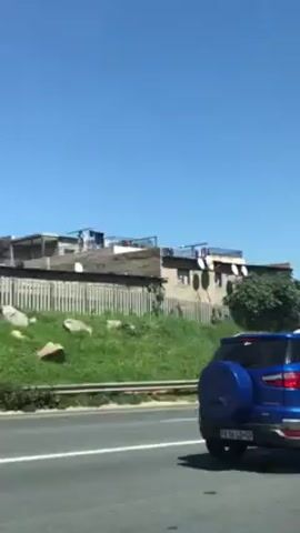The video shows a view from a moving car, looking at buildings with many satellite dishes along a highway. There are other cars on the highway, including a blue SUV and a silver station wagon. The sky is clear and blue. The buildings are a mix of brick and other materials, and some have fences around them. There's green vegetation along the highway divider.