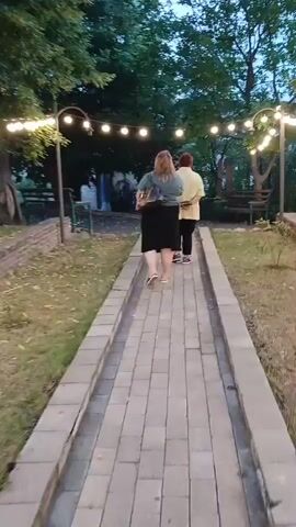 The video shows two women walking away from the camera on a paved pathway in an outdoor setting. The pathway is made of light-colored rectangular pavers and is bordered by low stone walls. To the left of the pathway, there is grass and some brickwork. To the right, there is dry grass and more low stone walls. String lights with round bulbs are strung overhead, illuminating the scene. The trees in the background are lush and green, suggesting it is daytime or early evening, likely warm weather. The sky appears a dusky blue, hinting at either early morning or late evening. The women are walking side-by-side, with one in front of the other. The woman in front is wearing a light-colored top and dark skirt, while the woman behind her is wearing a greenish-blue top and a dark skirt. They appear to be casually walking through an outdoor space, possibly a park, garden, or a venue. The overall mood is peaceful and relaxed. There is no visible text in the video. The location is identified as Telavi, Georgia.