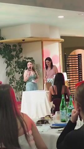 A woman in a sparkly teal dress stands at a white-draped table on a stage, holding a microphone and speaking. She gestures with her left hand. To her right, a woman in a pink top stands behind the table, looking towards the audience. A plant is visible on the left side of the stage. In the foreground, a woman with long dark hair is visible from the back, sitting at a table. Several green bottles of Perrier are on the table, along with other items. The background shows wooden blinds and what appears to be a wall. The lighting is warm, with some pinkish light spots visible on the stage.