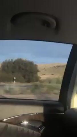 The video shows a view from inside a moving car, looking out the side window. The exterior environment is a rural, open landscape with a field and a distant, small hill. A small, derelict-looking building is visible in the field, set back from the road, as the car passes by. The sky is blue with some overcast areas. The car's interior is visible along the bottom of the frame, including the door panel with a wood grain accent. The overall impression is of a drive through a sparsely populated area.