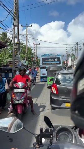 The media file captures a bustling street scene in Kodagoda, Sri Lanka, under a bright, partly cloudy sky, suggesting a warm, sunny day. The perspective is from a motorbike, looking forward into moderate traffic.

The primary subjects are various vehicles and people navigating a road lined with shops and utility poles adorned with numerous wires. A red scooter with a rider in a red shirt and a helmet is prominently in the foreground to the left. Further down the road, a grey Suzuki car is stopped, its license plate visible as "CAJ 9153". Behind it, a green and white tuk-tuk is also visible, along with a white and blue bus in the distance. Several other motorbikes and individuals are interspersed throughout the scene, some walking on the roadside.

The activity depicted is typical street traffic, with vehicles moving slowly and people going about their day. The presence of traffic lights and what appear to be railway crossing signals further indicate a busy intersection or route.

Notable details include the presence of many overhead electrical wires, characteristic of many South Asian countries. The shops lining the street display signs, including one that reads "SHIKAWAKA" and another with the brand "MEIZU". The overall mood appears to be one of everyday activity and movement, with no overt signs of distress or urgency, despite the traffic. The time of day is clearly daytime, given the strong sunlight and shadows.