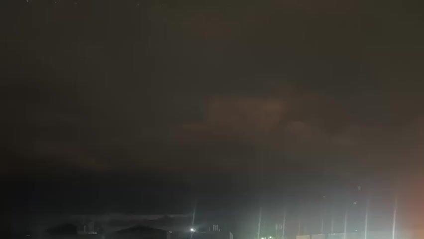 The video shows a dark, cloudy night sky with faint lights from buildings in the distance. Some of the lights appear to be streetlights or lights from within buildings, casting long vertical beams upwards. There are also some smaller, brighter lights in the sky that could be stars or distant aircraft. The clouds are thick and dark, obscuring any clear view of the sky above. The overall impression is of a somewhat gloomy and overcast night.