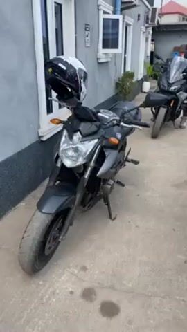 The video shows a collection of motorcycles parked outside a building with a light gray exterior. The bikes include a black Yamaha, a BMW adventure bike with saddlebags, a dark blue Yamaha MT-09, and several other motorcycles. Helmets are placed on the handlebars of some bikes. People wearing riding gear are present, and appear to be preparing to ride. The setting seems to be a paved area outside of a residential or commercial building.