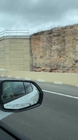 The video shows a car driving on a highway, with the view captured from the driver's side window, focusing on the side mirror. The mirror reflects a white car in the adjacent lane. The external environment consists of a beige, brick-patterned wall alongside the highway, which transitions into a rocky hillside. A yellow excavator is visible on top of the hillside. The sky is overcast and gray. Road markings, including a solid white line, are visible on the highway. The mirror's text, "OBJECTS IN MIRROR ARE CLOSER THAN THEY APPEAR," is clearly readable. The driver is using their phone, reflected in the side mirror.