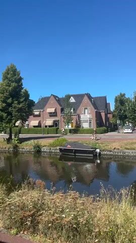 The video pans across a canal scene on a sunny day. In the foreground, there is lush green foliage and a red path. A dark gray lamppost is visible on the left. The canal itself is dark with reflections of the surroundings. On the other side of the canal, there is a walkway, a row of buildings, and trees. Several buildings of varying architectural styles are visible, including brick houses and modern structures. One building has awnings over its windows. There are trees lining the bank of the canal and behind the buildings. In the canal, a dark-colored boat is moored on the right side. People are seen cycling on the walkway. The sky is a clear, bright blue.