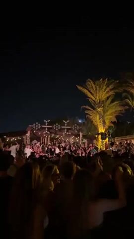 The video shows a crowded outdoor event at night. A large group of people are gathered, many of them dancing. In the background, there is a stage area with lights and decorations, possibly a DJ booth or performance setup. Palm trees are visible, suggesting a warm climate or tropical setting. The overall atmosphere appears to be festive and lively.