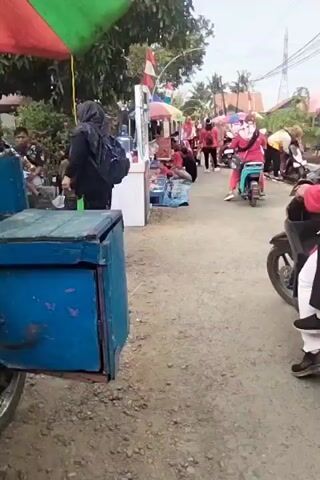 Two women on a motorcycle, the passenger in a pink hijab and the rider in a blue hijab, are positioned on a dirt road. The rider is wearing black pants and a blue top, while the passenger is wearing white pants and a pink top. They are facing away from the camera, towards a row of stalls or tents. To the left of the motorcycle, a blue cart is visible, with a person in a dark hijab standing behind it. In the background, several other individuals are present, some also on motorcycles, and the area appears to be an outdoor market or gathering. The overall environment suggests a warm, possibly humid climate, with trees and buildings visible in the distance, along with power lines.