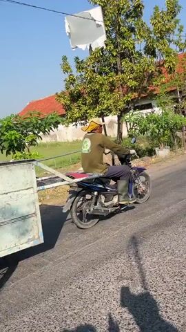 A man wearing a hat and green shirt rides a blue motorcycle pulling a trailer down a paved road. He is wearing green boots. The trailer is made of white metal. The man is looking forward. The road is next to a grassy area with trees and houses. The sky is blue and clear. There is a white sheet hanging from a pole in the background.