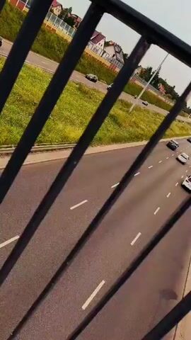The video shows a highway with cars driving on it, viewed from behind a railing. The highway is bordered by a grassy area with some trees and houses in the distance. The sky is cloudy. The camera is moving, creating a sense of motion. Several cars are visible on the highway, including a white van, a black car, and a white SUV. There are also some other vehicles further down the road. The perspective suggests the camera is on an overpass or a bridge. The overall atmosphere is mundane and ordinary, depicting a typical scene of traffic on a highway.