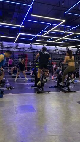 The video shows a group of people exercising in a gym. The gym has a modern design with blue and white neon lights on the ceiling. In the foreground, a man in a blue tank top is doing bicep curls with dumbbells. To his right, a woman in a black long-sleeved shirt and shorts is standing on a Bosu ball and performing an overhead press with a dumbbell. Further back, several other individuals are engaged in various exercises. Some are on Bosu balls, performing what appears to be a plank or a similar floor exercise. Others are on exercise bikes. The floor is a dark gray mat with white lines marking workout stations. The background features gym equipment like weight racks and exercise machines. The overall atmosphere suggests a group fitness class or a high-intensity workout session.