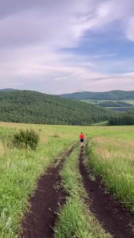 A person wearing an orange shirt and black shorts runs down a dirt path through a grassy field. The path is flanked by tall green grass. In the background, rolling hills covered in dense green forest stretch towards the horizon. The sky above is partly cloudy with patches of blue. The camera is positioned behind the runner, facing the direction they are running.