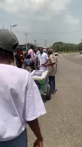 The video shows a group of people standing on the side of a road. Some are dressed casually, while others are wearing white shirts and black pants. There is a food vendor selling items from a cooler on wheels. The people appear to be interacting with each other. There is a police vehicle in the background. The weather appears sunny.