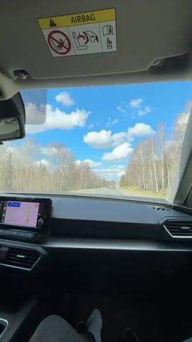 The video shows a first-person perspective from inside a car, looking out through the front windshield. The car is driving on a paved road that stretches ahead into the distance. On both sides of the road, there are numerous trees with white trunks, suggesting they might be birch trees, indicating a natural or forested environment. The sky above is bright blue with scattered white, fluffy clouds. Another car can be seen driving ahead in the distance.

Inside the car, parts of the dashboard, air vents, and an infotainment screen displaying a navigation map are visible. The top of the frame shows the car's sun visor, which has "AIRBAG" written on it along with several safety icons related to airbag operation and child seat placement. The legs and feet of a person, presumably the driver or a passenger, are visible in the lower part of the frame. The time of day appears to be bright and sunny. Background audio includes music and faint voices. The location context is Pechmen, Russia.