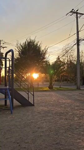 The video shows a playground at sunset. A black slide is in the foreground on the left. The sun shines brightly through trees in the center of the frame. Behind the trees, on the street, are cars and a person walking a dog. The person is silhouetted by the sunlight. The ground of the playground is tan dirt. Power lines are visible in the sky.