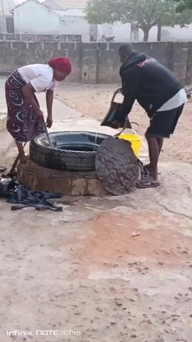Two individuals are engaged in what appears to be well maintenance or cleaning activities in an outdoor setting. A woman, wearing a white top, a patterned skirt, and a red head covering, is using a tool to manipulate something inside a tire that forms part of the well structure. A man, clad in a black hoodie, a white undershirt, and black shorts, is lifting a black bag or container, possibly filled with debris, out of the well. A yellow bucket and a metallic lid or cover are positioned nearby. The well itself is made of tires stacked on a concrete base. Several black rubber straps or materials are lying on the ground near the well. The surrounding environment includes a cement or stone-paved area, a brick wall, and a few buildings and trees in the background. The sky appears bright, suggesting it's daytime. The video's label indicates it was taken using an Infinix NOTE 30 Pro.