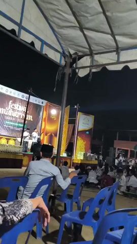 The video shows an outdoor night event under a large tent with blue plastic chairs in the foreground. In the background, a stage is set up with banners. One banner displays "Festival Muharram 1445 H 2023 M". Several people are on the stage, and a large crowd is seated on the ground in front of it, many of whom appear to be young males wearing white attire and caps. The atmosphere suggests a religious or cultural gathering. The lighting is artificial, illuminating the stage and the audience area.