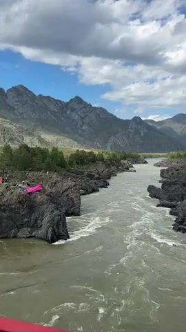 The video shows a wide, fast-flowing river with greyish-green water. The river is flanked by rocky cliffs covered with some trees.  A bright pink kayak sits on the rocks on the left side of the frame, near a small group of people.  In the background, towering mountains are visible, partially obscured by clouds in the blue sky. The weather appears partly cloudy with patches of blue sky.  The camera seems to be positioned on a bridge, offering a high-angle view of the scene.