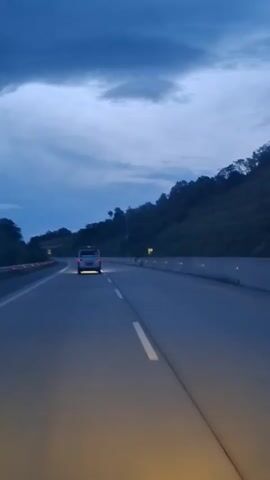 The video shows a view from inside a vehicle driving on a highway at dusk. A white van is visible ahead, driving in the same direction. The sky is dark blue with some clouds. The highway has multiple lanes, and there are trees and hills on the right side of the road. The headlights of the vehicle from which the video is recorded illuminate the road ahead.